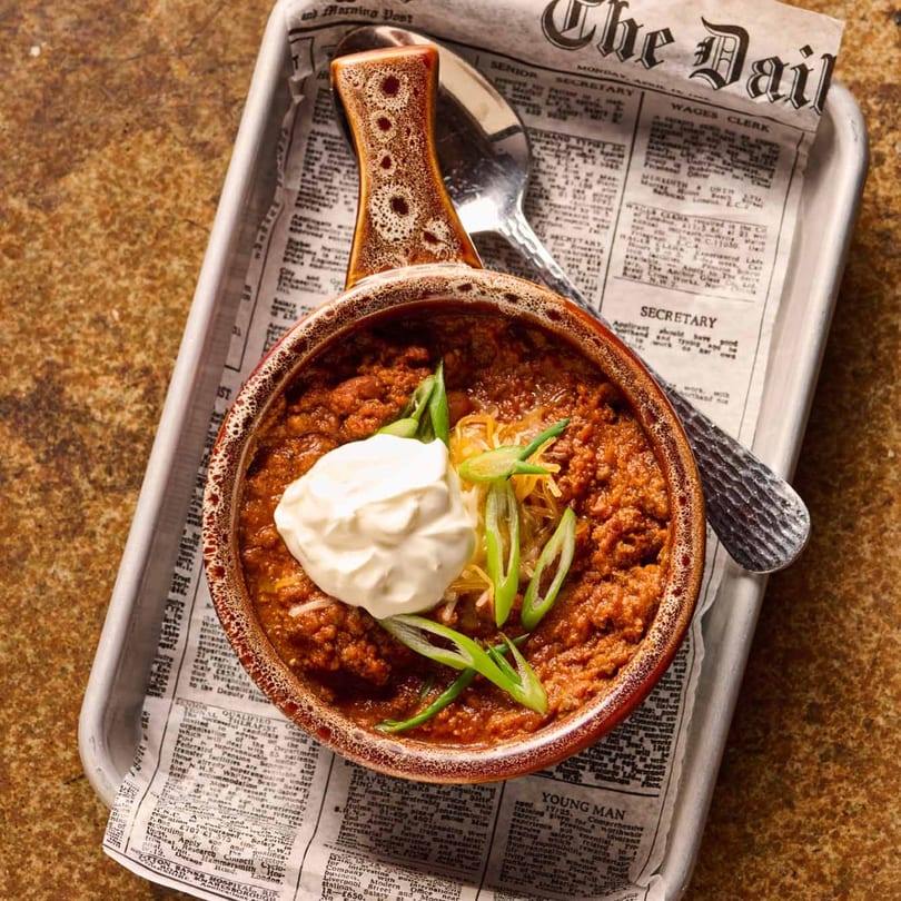 A bowl of chili topped with sour cream, shredded cheese, and green onions, served on a newspaper-lined tray.