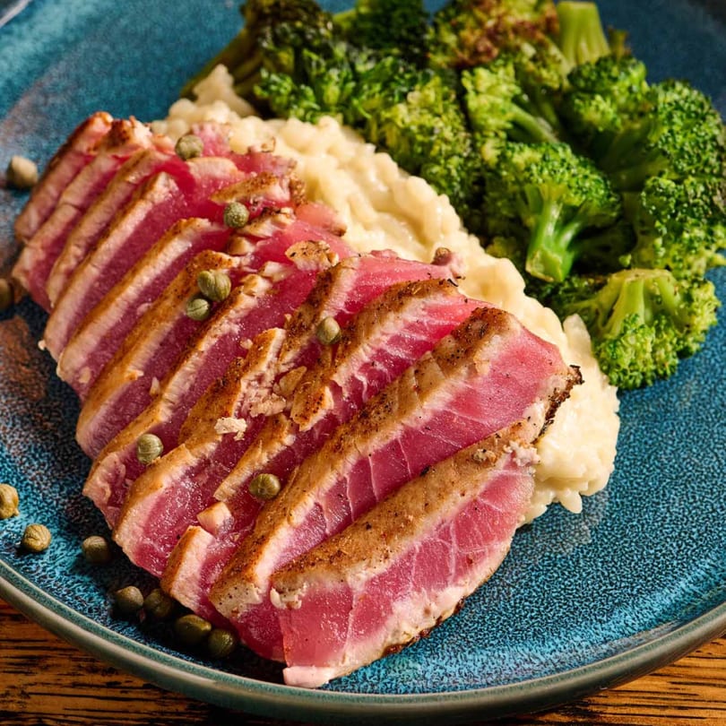 Seared tuna slices with capers, creamy risotto, and steamed broccoli on a blue plate.