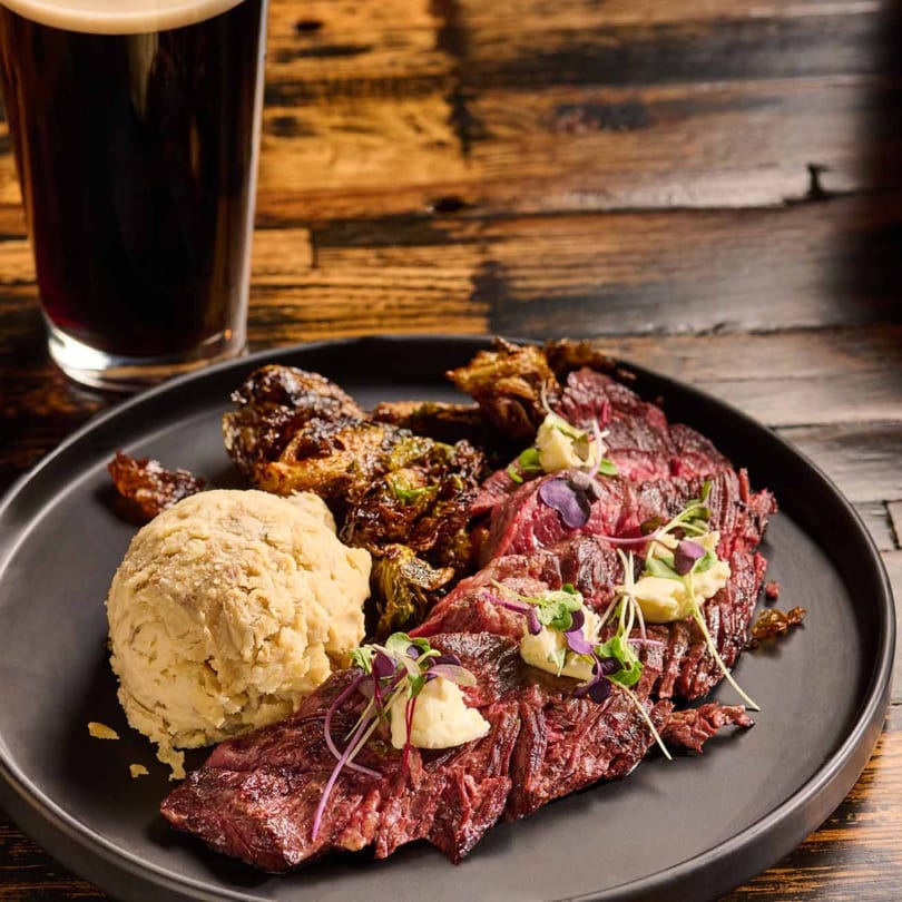 Grilled steak with mashed potatoes, roasted Brussels sprouts, and a dark beer on a wooden table.
